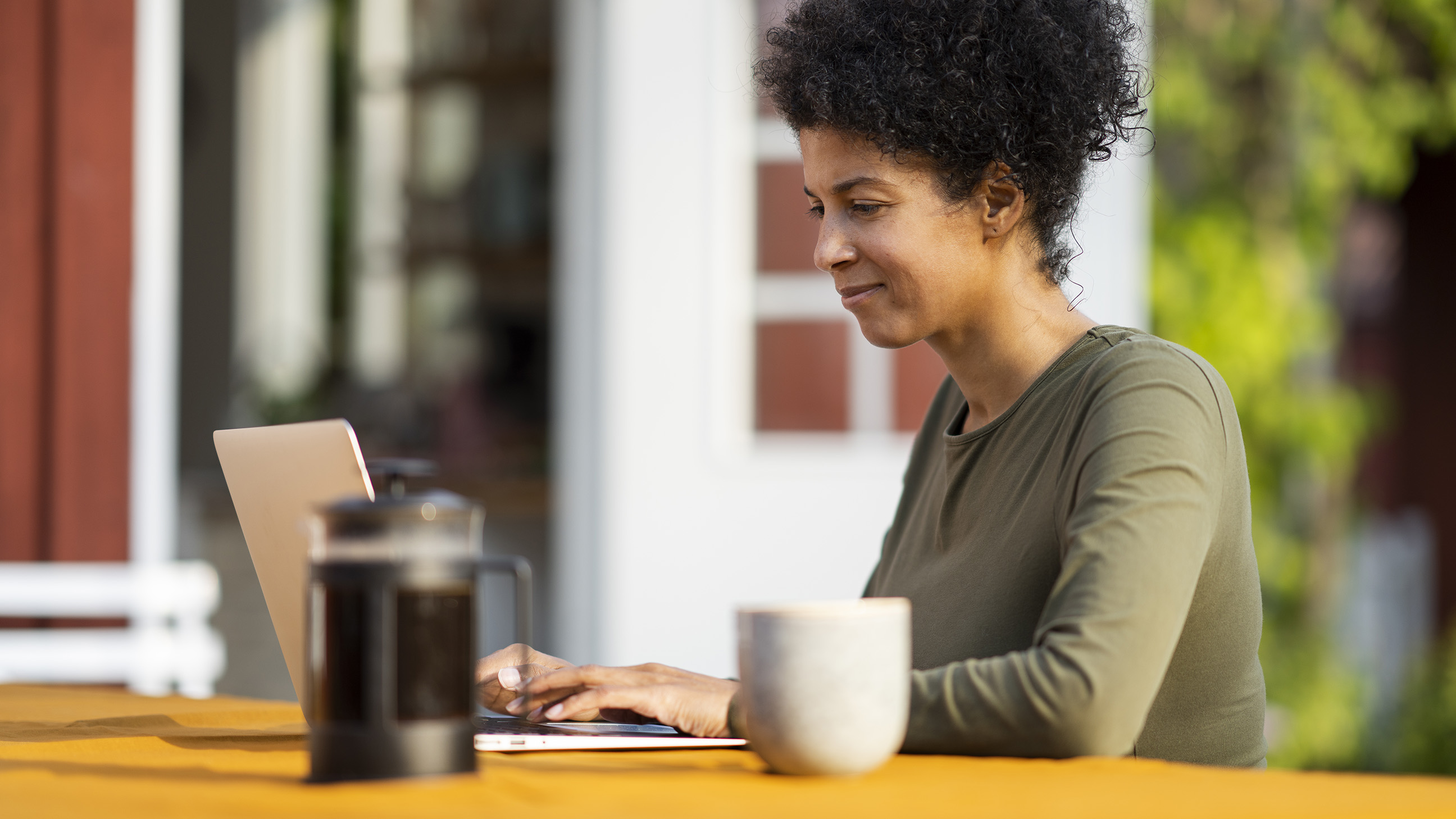 En kvinna sitter utomhus framför sin laptop och dricker kaffe. 